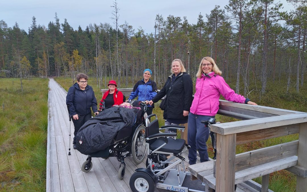 Kuusi aikuista ihmistä on puisella pitkospuulla suomaisemassa. Osa käyttää apuvälineitä, ja etualalla on sähköinen pyörätuoli. Pitkospuita reunustaa harva mäntymetsä, ja taustalla näkyy kosteikkoa ja puita.