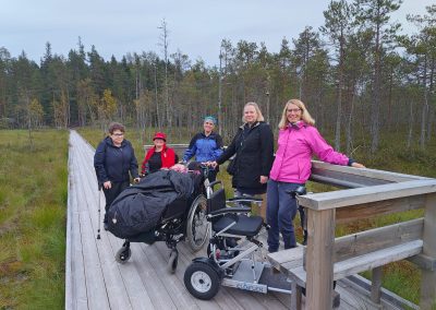 Kuusi aikuista ihmistä on puisella pitkospuulla suomaisemassa. Osa käyttää apuvälineitä, ja etualalla on sähköinen pyörätuoli. Pitkospuita reunustaa harva mäntymetsä, ja taustalla näkyy kosteikkoa ja puita.