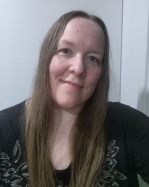 Hymyilevä lähikuva Outista valkoisen seinän edessä. Outilla on pitkät vaaleanruskeat hiukset ja tummat vaatteet.