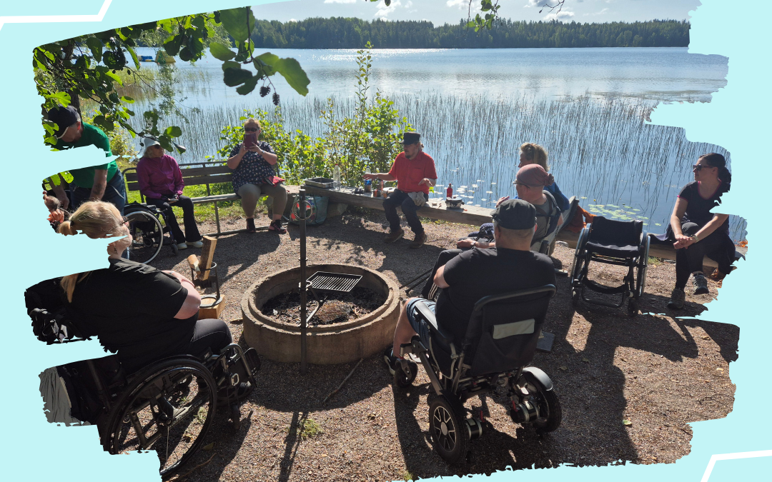 Ryhmä ihmisiä istuu ulkona järven rannalla nuotion ympärillä. Useilla osallistujilla on pyörätuoli tai sähköpyörätuoli. Taustalla näkyy järvi, ruovikkoa ja metsää aurinkoisessa säässä.