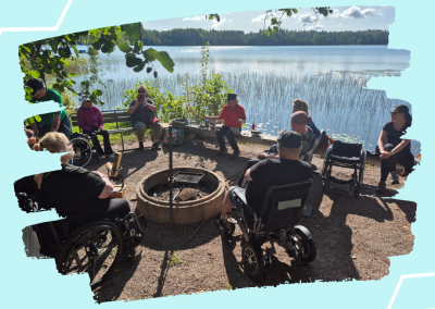 Ryhmä ihmisiä istuu ulkona järven rannalla nuotion ympärillä. Useilla osallistujilla on pyörätuoli tai sähköpyörätuoli. Taustalla näkyy järvi, ruovikkoa ja metsää aurinkoisessa säässä.