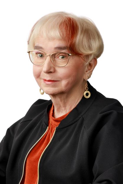 Vuokko Klasilan kuva vaaleataustaa vasten. Vuokko istuu hieman sivuttain kameraan päin ja katsoo suoraan eteenpäin. Vuokolla on lyhyt vaalea tukka, jossa on punertava sävy otsan kohdalla. Henkilö käyttää kapeita silmälaseja, mustaa takkia ja oranssia paitaa. Korvissa on roikkuvat, pyöreät korvakorut. Valaistus on tasainen ja kuva on rajattu rintakehän yläosasta.