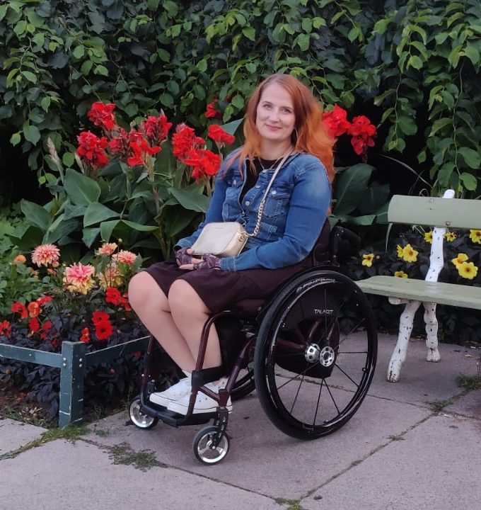 Pyörätuolia käyttävä henkilö istuu puiston kävelyreitin varrella kukkaistutusten edessä. Taustalla on punaisia ja vaaleita kukkia sekä vihreää kasvillisuutta. Henkilöllä on farkkutakki, tumma mekko ja vaalea olkalaukku. Vieressä näkyy vaalea puistonpenkki. Kuvassa on kesäinen ja vehreä puistomaisema.